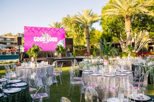 Outdoor event setup with a stage featuring a large screen displaying the text "GOOD GOOD". Round tables with silver sequin tablecloths and clear chairs are arranged on a green lawn. Palm trees and a pool can be seen in the background.