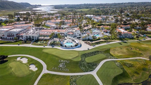 Aerial view of Omni La Costa Resort & Spa, showcasing a luxurious resort with golf courses, multiple pools, white buildings, and a lush landscape with palm trees and a lake in the background.